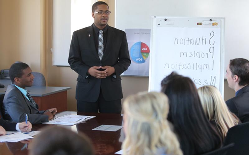 man stands in front of group at a conference table and presents information on white board