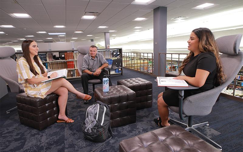 three peopel sitting in chairs in a libary facing eachother having a discussion