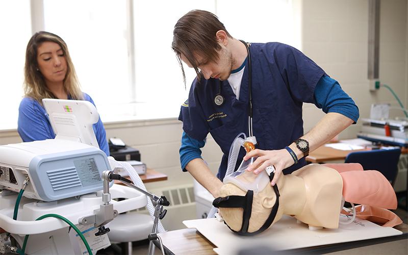 A man using medical tools to practice repiratory care on practice dummy wile being supervised by another woman